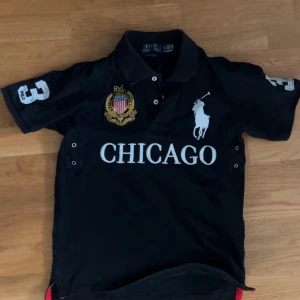 Svart Polo Ralph Lauren t-shirt Chicago - Svart t-shirt från Polo Ralph Lauren med stor vit CHICAGO-text och logga på bröstet, broderad emblem och nummer 3 på både ärm och rygg. Klassisk pikékrage och knappslå framtill. Perfekt för dig som gillar sportig och preppy stil.kan gå ner i pris vid snabbaffär