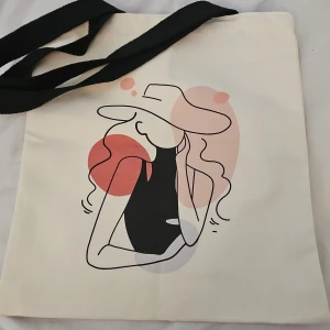 Beige tygkasse med konstmotiv - Snygg beige shoppingväska i bomull med svart axelrem. Framsidan har ett stilrent konstnärligt tryck av en tjej med hatt i svart, rött och rosa toner. Perfekt för att bära böcker, laptop eller shopping. Enkel och trendig design som sticker ut. Fick den i present för något år sedan, vet tyvärr inte märket. Aldrig använd men lite lite smutsig då den legat i min garderob och samlat lite damm, men inget som syns mycket.