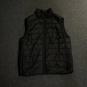 Svart dunväst i S - Svart dunväst med ståkrage och horisontella sömmar. Västen har dragkedja framtill och är tillverkad i ett glansigt syntetmaterial. Beställ gärna med bring då kan jag leverera mycket snabbare.priset kan diskuteras.