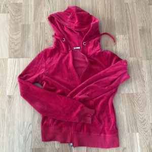 Röd velour hoodie med dragkedja - Säljer en röd hoodie i mjuk velour med huva och snörning. Tröjan har långa ärmar, ribbade muddar och praktiska fickor framtill. Dragkedja hela vägen och coolt metallspänne. Perfekt för chill dagar och när du vill vara extra comfy.