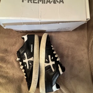 Snygga svarta sneakers från Premiata - Snygga svarta sneakers från Premiata med vita detaljer och logga på sidan. Skorna har vita snören, svart ovandel i skinn och vit sula med mönster. Perfekta för dig som gillar stilrena och trendiga skor med en sportig vibe.