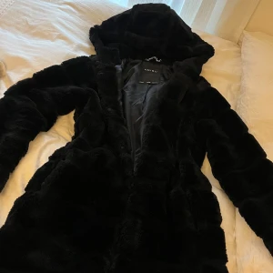 Svart pälsjacka med luva från new yorker - Mysig svart pälsjacka från newyorker med luva och långa ärmar. Jackan är fluffig och har en snygg, lite oversized passform. Perfekt för kalla dagar när du vill vara både varm och stilren.