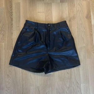 Svarta skinnshorts från Stradivarius - Snygga svarta shorts i skinnimitation från Stradivarius med hög midja och bälteshällor. De har knapp och dragkedja framtill, samt fickor både fram och bak. Perfekt med ett par strumpbyxor och en stickade tröja 💗