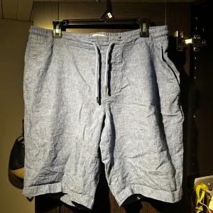 Blå linneshorts med snörning - Snygga blå shorts i linne med avslappnad passform och elastisk midja med snörning. Shortsen har sidofickor fram och två bakfickor med knappar