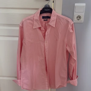 Rosa skjorta från Polo Ralph Lauren - Säljer en klassisk rosa skjorta från Polo Ralph Lauren i storlek L. Skjortan är i bomull, har lång ärm, knappar framtill och en snygg custom fit-passform.  Använd 2 gånger, köpt på nk i Stockholm. 