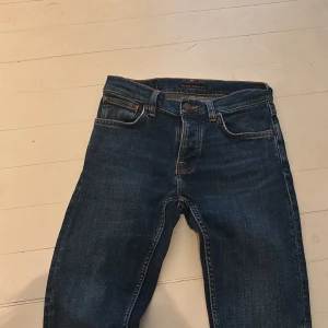 Nudie Jeans blå jeans 28/30 - Snygga blå jeans från Nudie Jeans med klassisk femficksdesign och ikoniska broderade bakfickor. Jeansen har orangea kontrastsömmar och är tillverkade i slitstarkt denim. Perfekt passform och stilren look för dig som gillar tidlösa jeans. modell grim tim 