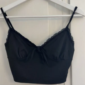 Svart croppad linnetopp med spets - Snygg svart croppad linnetopp med tunna axelband, spetsdetaljer längs kanterna och en liten rosett framtill. Perfekt att styla med jeans eller kjol för en trendig look.