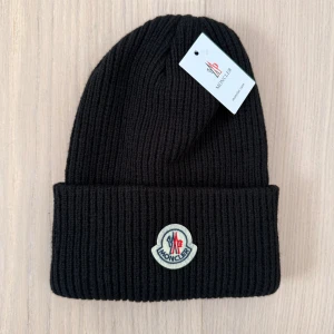Svart ribbad Moncler mössa - Svart ribbstickad mössa från Moncler med uppvikt kant och klassisk Moncler-logga framtill. Snygg och enkel design som passar till allt, tillverkad i mjuk ull som håller dig varm. Perfekt accessoar för kalla dagar och en stilren look. Men den var för liten 