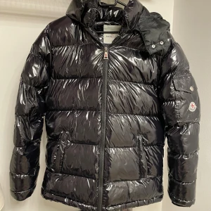 Moncler vinterjacka - Snygg svart pufferjacka från Moncler med glansig finish, huva och dragkedja framtill. Jackan har Moncler-logga på ärmen och stora fickor. Perfekt för kalla dagar när du vill hålla stilen och värmen.