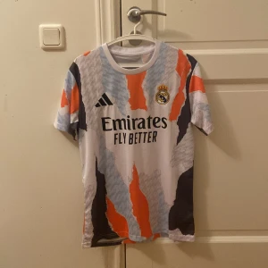 Real Madrid x Adidas fotbollströja M - Säljer en Real Madrid fotbollströja från Adidas i slim fit. Tröjan har korta ärmar och ett unikt abstrakt mönster i vitt, svart, blått och orange. Materialet är lätt och andas, perfekt för träning eller match. Emirates Fly Better-logga på bröstet.