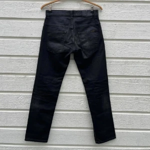 Svarta jeans från Nudie Jeans - Snygga svarta jeans från Nudie Jeans med klassisk femficksdesign och raka ben. Jeansen har en mörk tvätt och subtila slitningar för en cool look. Tillverkade i slitstarkt jeanstyg med knappstängning och bälteshällor.