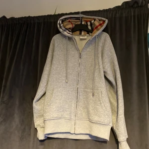 Grå Burberry hoodie med rutig huva - Grå hoodie från Burberry med klassisk rutig insida på huvan. Tröjan har dragkedja framtill, snörning i huvan och två fickor. Ikoniska Burberry-detaljer på dragkedja och snörändar. Perfekt för dig som vill ha en stilren och lyxig look.