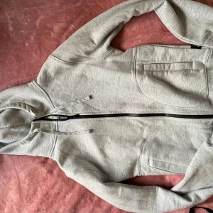 Grå hoodie med dragkedja och tumhål - Snygg ljusgrå hoodie med hel dragkedja framtill, huva med snörning och praktiska tumhål i ärmsluten. Tröjan har två fickor på sidorna och är tillverkad i mjuk bomullsmix. Perfekt för chill eller när du är på språng.