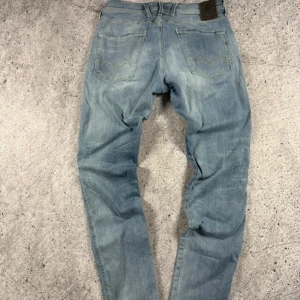 Replay Anbass ljusblå jeans - Snygga ljusblå Replay Anbass jeans i stretchigt Hyperflex-material. Klassisk femficksmodell med raka ben och diskreta slitningar. Märkespatch bak i midjan och broderad detalj på bakfickan. Perfekta för en avslappnad streetstil.