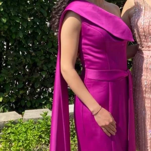 Lila långklänning med släp och bälte - Säljer en elegant lila långklänning med figurnära passform, ett axelbandslöst snitt och ett brett omlottliknande tyg över axeln. Klänningen har ett matchande bälte i midjan och ett långt släp som ger extra drama. Perfekt för bal eller festliga tillfällen. 34/36