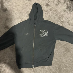 Grå  hoodie Supply & Demand - Grå hoodie med dragkedja från Supply & Demand storlek XS. Tryck på bröstet med texten 'Born in Brooklyn Raised on the Streets' och en cool grafisk logga. Klassisk huva, känguruficka och mjukt material. Perfekt streetstyle-vibe. Jätte bra skick har aldrig använt pga för liten storlek.