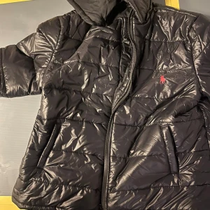 Svart pufferjacka  - Svart pufferjacka från RL med glansig finish och klassisk röd logga på bröstet. Jackan har huva, dragkedja framtill och två fickor. Perfekt för kalla dagar och riktigt snygg till streetwear-stilen.