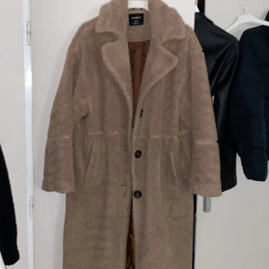 Beige teddyjacka från Lindex - Mysig beige teddyjacka från Lindex med stora knappar framtill och bred krage. Jackan är lång, har två fickor och är fodrad med brunt tyg på insidan. Perfekt för kalla dagar när du vill vara både varm och snygg.