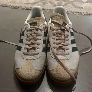 Adidas Gazelle sneakers vit/grön mocka - Säljer ett par Adidas Gazelle sneakers i vit mocka med gröna detaljer och klassiska tre ränder på sidan. Skorna har beige snörning, gummisula och grönt parti vid hälen. Tungan är i ruggad mocka och insidan är vit. Perfekta för dig som gillar retrostil och streetwear.