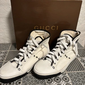 Gucci vita high-top sneakers - Snygga vita high-top sneakers från Gucci med svarta detaljer och svart sula. Skorna har snörning, rund tå och är tillverkade i en mix av skinn och textil. Skon och lådan är original, cirka 15 år gammal. Köpt på NK, prislapp kvar på låda.snören i original (svarta) ingår samt ett par helt nya Gucci-snöre. Canvasen är behandlad och sträv på utsidan. Snören är nya (vita) sulan är behandlad med sneakerfärg. Låda, påse, snöre i original ingår. 
