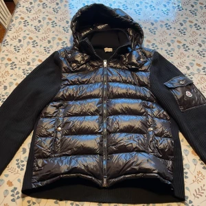 Svart pufferjacka från Moncler - Snygg svart pufferjacka från Moncler med glansig dunfylld front, stickade ärmar och huva. Jackan har dragkedja, fickor med tryckknappar och Moncler-logga på ärmen. Perfekt för dig som vill ha en stilren och exklusiv look under kyliga dagar.
