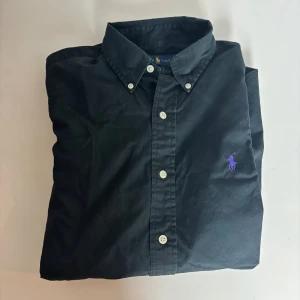 Svart skjorta från Ralph Lauren S - Klassisk svart skjorta från Ralph Lauren med lila broderad logga på bröstet. Skjortan har lång ärm, knappar framtill och button-down krage. Tillverkad i mjuk bomull som känns skön mot huden. Perfekt för dig som gillar stilrena och tidlösa plagg.