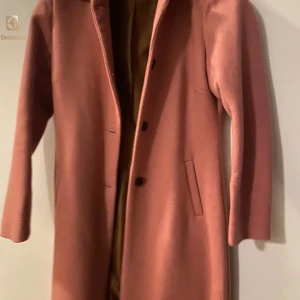 Rosa ullkappa med knappar - Snygg rosa kappa i ull med klassisk rak passform och diskreta knappar framtill. Jackan har två fickor på sidorna och är helfodrad. Perfekt för dig som vill ha en stilren och trendig look i höst och vinter. 