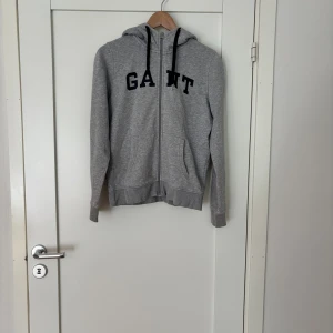 Grå hoodie från GANT med dragkedja - Grå hoodie från GANT med svart tryck över bröstet och hel dragkedja framtill. Tröjan har huva med svarta snören, två fickor och ribbade muddar vid ärmslut och nederkant. Perfekt för chill dagar eller när du vill ha en clean streetstyle.