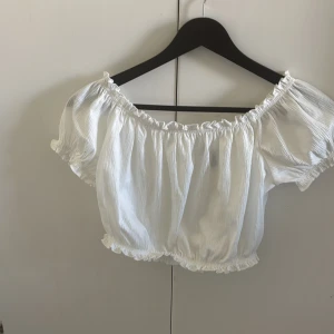 Vit offshoulder croppad topp - Säljer en söt vit croppad topp med offshoulder-modell och korta puffärmar. Toppen har resår vid axlar och midja samt små volangdetaljer längs kanterna. Perfekt för en somrig look!