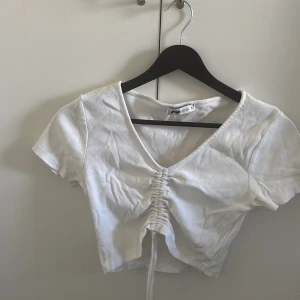 Vit croppad topp med snörning - Säljer en vit croppad topp från New Yorker med korta ärmar och v-ringning. Toppen har snygg snörning framtill som ger en cool rynkad effekt. Perfekt till jeans eller kjol för en trendig look.