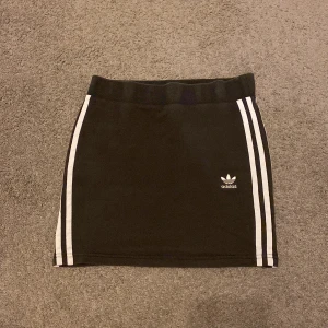 Svart kortkjol från Adidas med ränder - Svart kortkjol från Adidas med klassiska vita ränder längs sidorna och Adidas-logga framtill. Kjolen har en elastisk midja och är gjord i mjuk bomullsblandning. Sportig och stilren modell som är enkel att matcha.