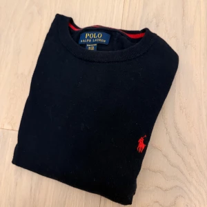Svart tröja från Polo Ralph Lauren XL - Klassisk svart tröja från Polo Ralph Lauren i mjuk bomull. Tröjan har rund halsringning med röd detalj på insidan och den ikoniska röda broderade loggan på bröstet. Perfekt för dig som gillar stilren och tidlös design.