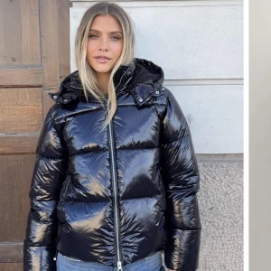 Svart glansig pufferjacka med huva - Säljer en svart glansig pufferjacka med huva och dragkedja framtill. Jackan har en quiltad look och är perfekt för kalla dagar. super snygg och lite fluffig inuti 
