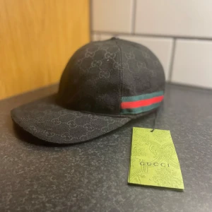 Svart keps från Gucci med logomönster - Svart keps från Gucci med diskret GG-logomönster över hela ytan. Klassisk böjd skärm och ikoniskt grönt och rött band på sidan. Kepsen är perfekt för dig som vill sticka ut  obs! 