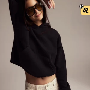 Svart croppad hoodie med huva - Svart croppad hoodie med huva och lång ärm. Tröjan har en loose passform och är tillverkad i mjukt material, perfekt för en chill och avslappnad stil. Klassisk känguruficka framtill och ribbade muddar vid ärmslut och nederkant.