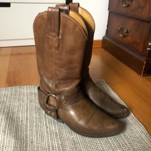 Bruna cowboy boots från Tony Mora - Snygga bruna cowboy boots från Tony Mora i äkta skinn. Klassisk westernstil med rund tå, dekorativa sömmar och metallspänne vid ankeln. Perfekta för dig som vill ha en cool och tidlös look med boots som sticker ut. Väldigt vintage. Säljer då dom är för smala för mig. 