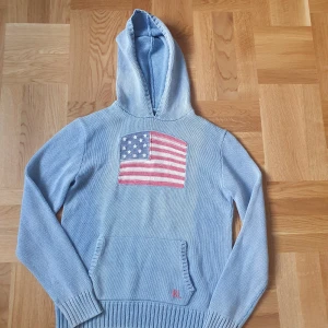 Ljusblå stickad hoodie från Ralph Lauren - Snygg ljusblå stickad hoodie från Ralph Lauren med amerikansk flagga framtill och RL-broderi på fickan. Tröjan har huva, känguruficka och långa ärmar. Perfekt för dig som vill ha en chill och stilren look. Det står att den är xl i barnstorlek vilket motsvarar s/m