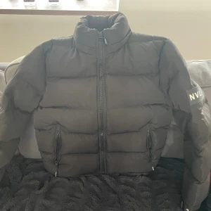 Svart pufferjacka från NVLT - Säljer en svart pufferjacka från NVLT med hög krage och dragkedja framtill. Jackan har två fickor med dragkedja och en patch med NVLT-logga på ena ärmen. Perfekt för kalla dagar och riktigt snygg till streetwear-stilen.