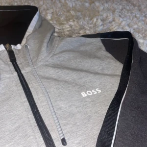 Grå och svart hoodie från BOSS - Snygg hoodie från BOSS i grått och svart med vit logga på bröstet. Tröjan har hel dragkedja, huva med dragsko och svarta detaljer längs ärmarna. Perfekt för en chill och sportig look.