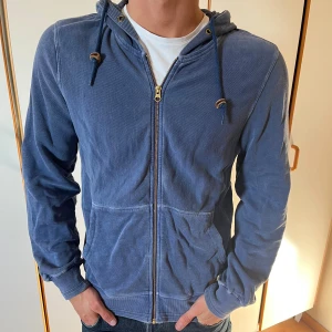 Blå hoodie från Woolrich med dragkedja - Säljer en blå hoodie från Woolrich i storlek M. Tröjan har huva med snörning, dragkedja framtill och två stora fickor. Tillverkad i mjuk bomull och har ribbade muddar vid ärmslut och nederkant. Perfekt för chill dagar eller när du vill ha en avslappnad stil. Passar S-M