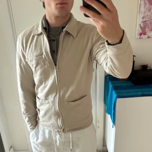 Beige overshirt från Riley  - Stilren beige overshirt från Riley. Jackan har klassisk krage, dragkedja framtill och tre fickor. Perfekt för lager-på-lager och enkel att matcha med olika outfits. Snygg och avslappnad passform med lång ärm. Bra skick.