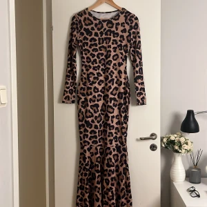 Lång leopardmönstrad klänning i mermaid passform - Snygg långärmad klänning med leopardmönster i brun/beige och svart. Klänningen är figurnära upptill och har en lätt utsvängd nederdel som ger en elegant siluett. Perfekt för dig som vill sticka ut med ett djurmönster och en trendig look.