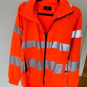 Orange vindjacka med reflexdetaljer XS - Säljer en orange vindjacka i storlek XS med breda reflexränder runt ärmar och kropp. Jackan har dragkedja framtill, hög krage och två fickor med dragkedja. Perfekt för att synas i mörkret och har en sportig vibe.
