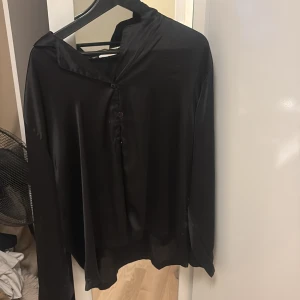 Svart silkeslen henley-skjorta - Svart henley-skjorta i silkeslent material med lång ärm och knappar framtill. Skjortan har en avslappnad passform och en lätt glansig yta som ger en stilren och modern look. Perfekt för dig som gillar minimalistisk stil. Oversize!