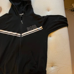 Svart hoodie från Hugo Boss - Svart hoodie från Hugo Boss med vit logga och två vita ränder över bröstet. Tröjan har dragkedja framtill, huva och fickor. Perfekt för en chill och stilren look.