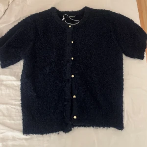 Mörkblå kofta från Gina Tricot - Säljer en fluffig mörkblå kofta från Gina Tricot med korta ärmar och snygga guldfärgade knappar framtill. Koftan har en mysig, lite lurvig textur och rund halsringning. Perfekt för lager på lager under kyliga dagar.