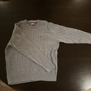 Ljusblå kabelstickad tröja Gant - Säljer en ljusblå kabelstickad tröja från Gant med rund halsringning och diskret logga broderad på bröstet. Tröjan har långa ärmar och ribbade muddar vid ärmslut och nederkant. Perfekt för dig som gillar klassisk och stilren look.strl 164 13-14 år