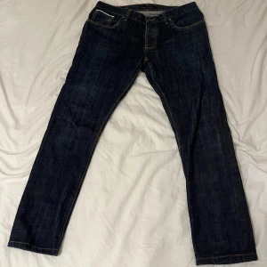 Mörkblå jeans från Nudie Jeans - Säljer ett par mörkblå jeans från Nudie Jeans med klassisk femficksdesign och kontrastsömmar. Jeansen har rak passform och är tillverkade i slitstarkt denimtyg av bomull. Snygga detaljer med Nudie-logga på bakfickan och läderpatch i midjan. Byxan är i w36 L34 men jag har w34L34 och passar mig perfekt så skulle säga att dem är i den storleken 