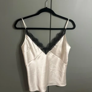 Vit satinlinnetopp med spets Gina Tricot - Supersnygg vit linnetopp i satin från Gina Tricot med tunna axelband och v-ringning dekorerad med svart spets. Toppen har en slät, glansig yta och justerbara axelband för perfekt passform. Perfekt att styla med jeans eller kjol för en trendig look.