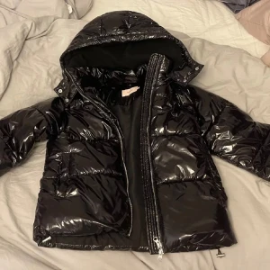 Trendig Svart glansig pufferjacka från Nelly  - Säljer en svart Nelly pufferjacka med glansig finish och stor huva. Jackan har dragkedja framtill, två stora fickor och quiltad design. Perfekt för kalla dagar när du vill hålla stilen och värmen. Priset går att diskutera.